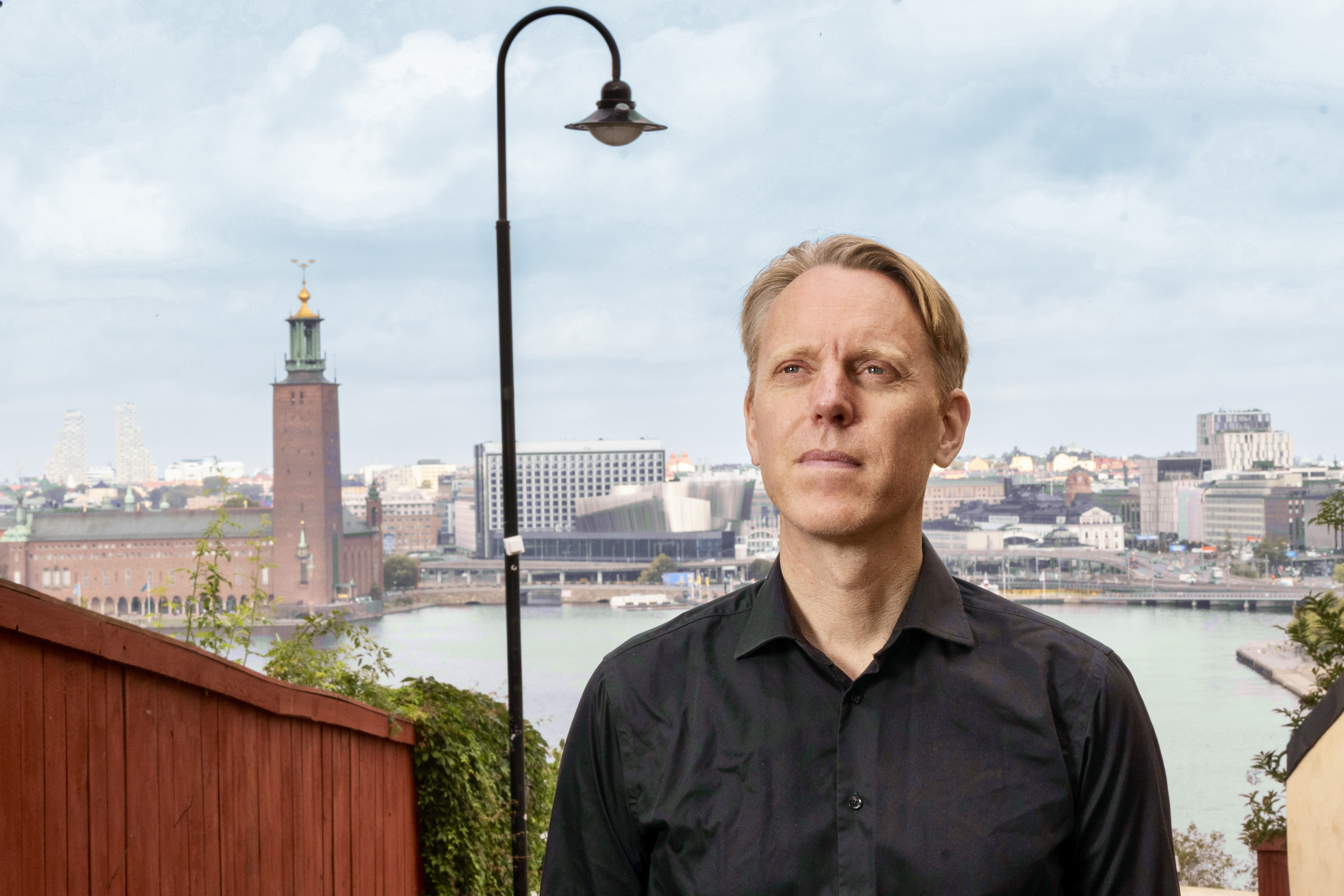 En man i svart skjorta står utomhus med en stad, en flod och ett torn i bakgrunden under en delvis molnig himmel.