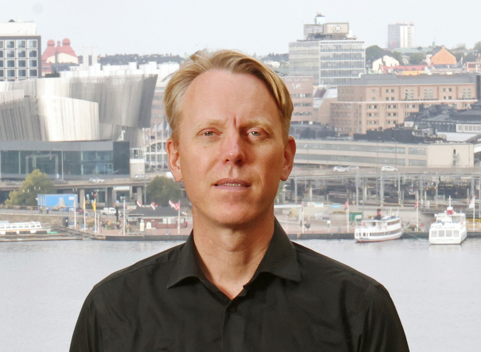 En man med kort blont hår och svart skjorta står utomhus framför en stad vid vattnet med byggnader och båtar i bakgrunden.