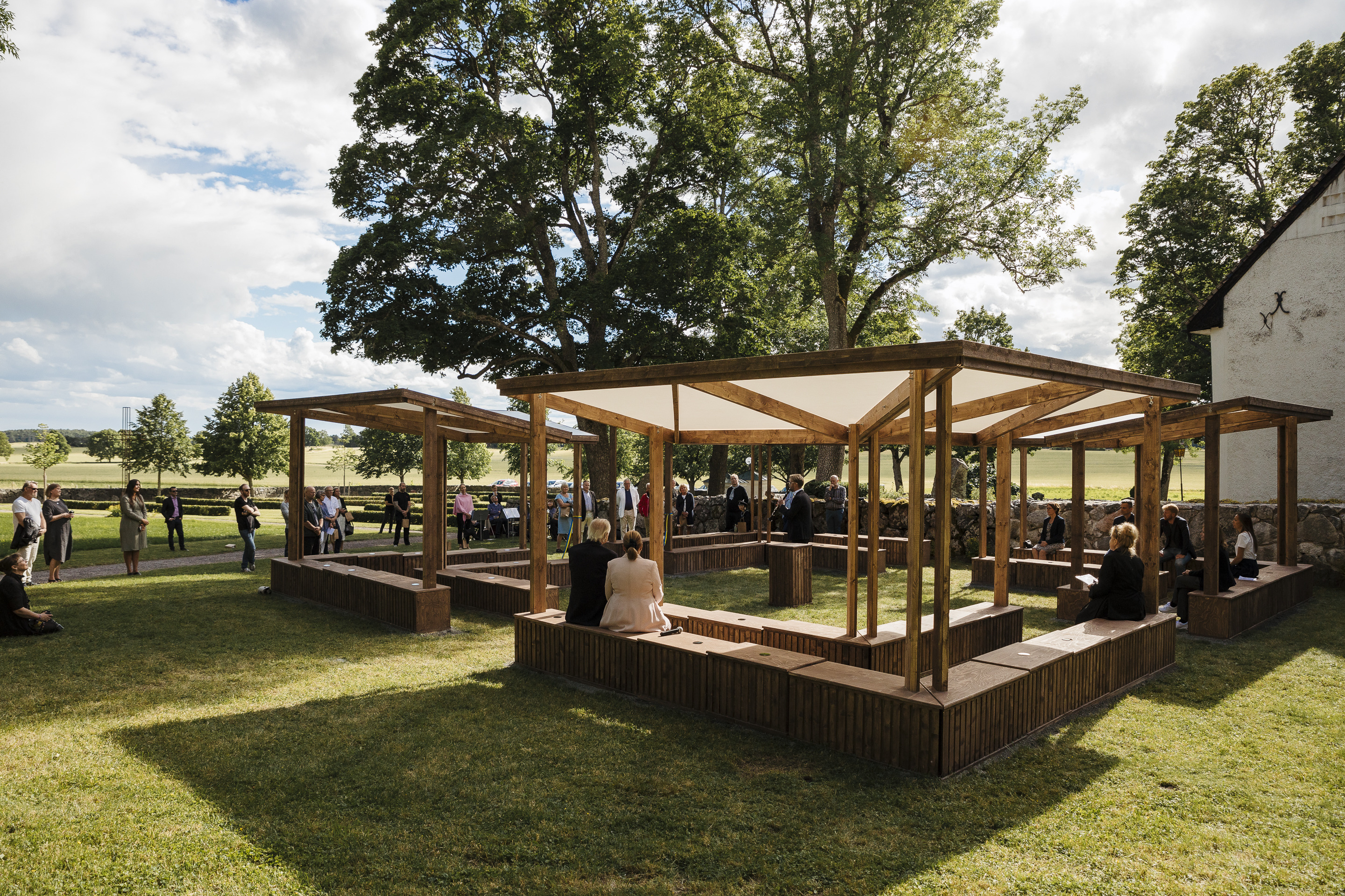 En grupp personer sitter och står runt träbänkar under öppna träskydd på en gräsmatta med träd och öppna fält i bakgrunden.