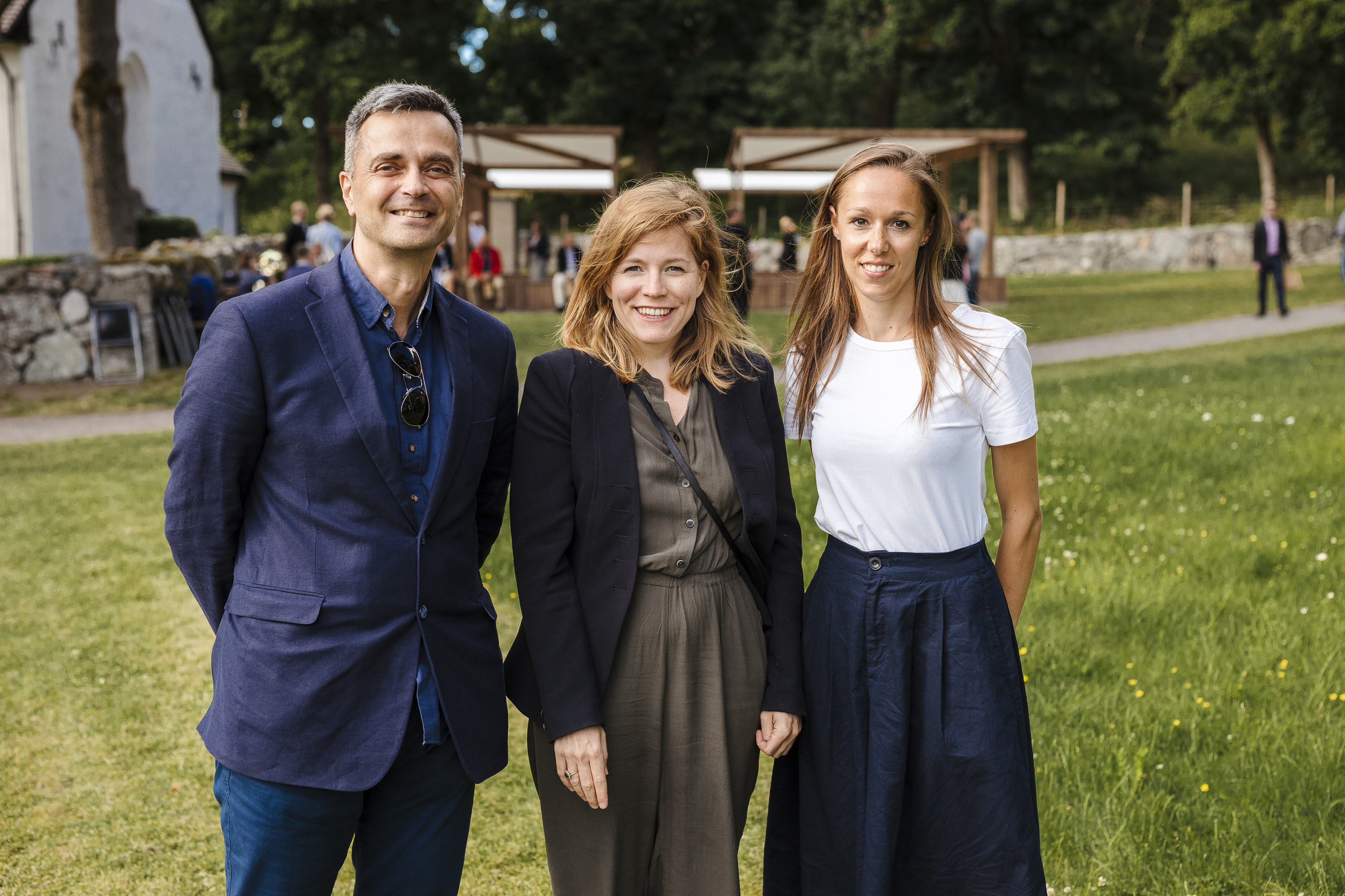 Tre personer står tillsammans utomhus på gräs, klädda i vardagliga och formella kläder, med träd och byggnader i bakgrunden.