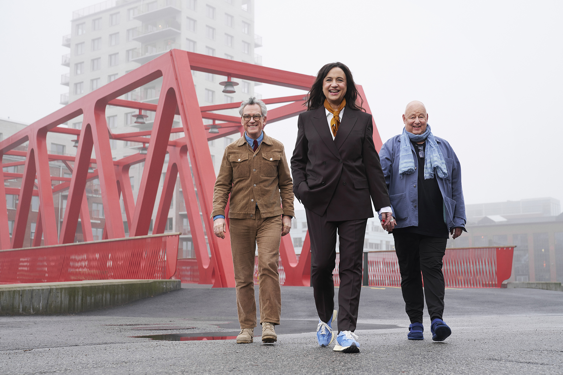 Tre personer går tillsammans på en stadsbro med rött räcke och moderna byggnader i bakgrunden en dimmig dag.