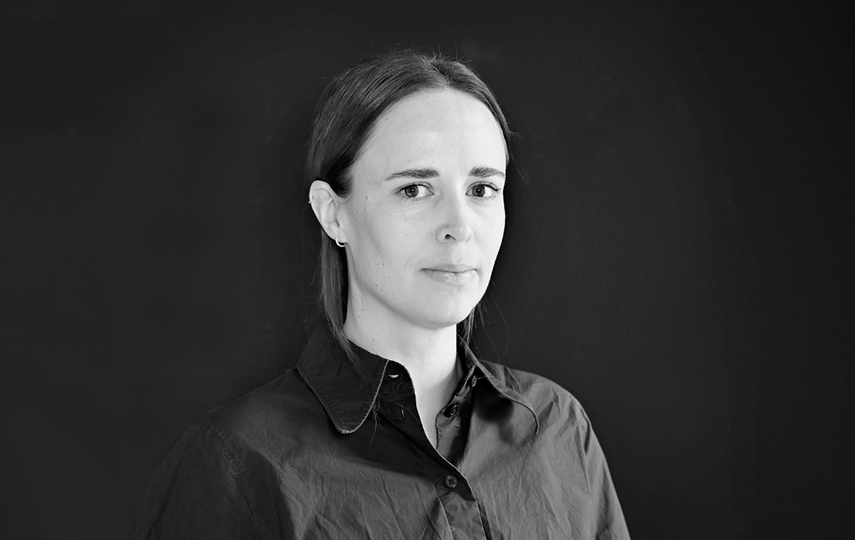 En kvinna med rakt brunt hår och neutral min står mot en mörk bakgrund, iklädd en knäppt mörk skjorta med krage.