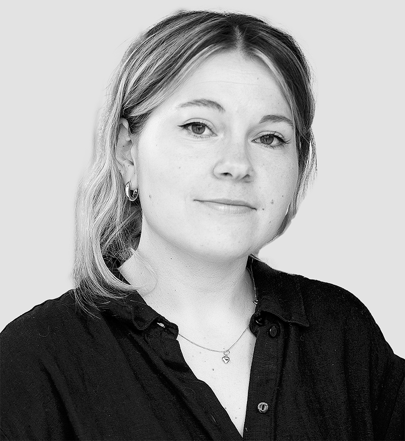 En kvinna med ljust hår, iförd en mörk skjorta med knappar och ett halsband, tittar mot kameran framför en ljus bakgrund.