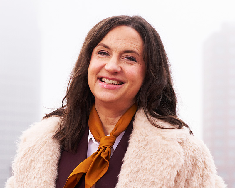 En kvinna med långt brunt hår har på sig en beige pälsjacka, en brun halsduk och en vit blus, utomhus framför byggnader.