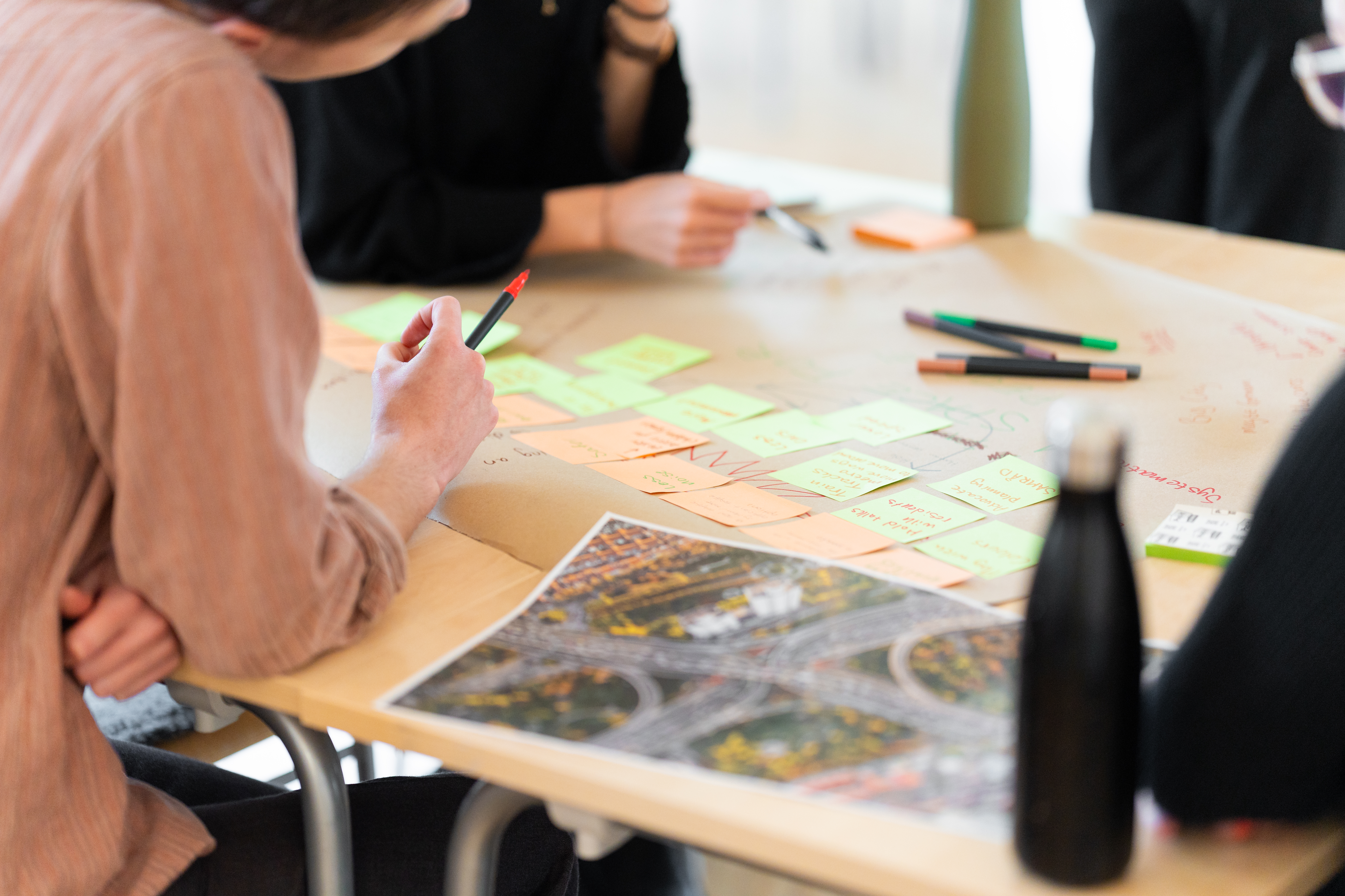 Flera personer sitter runt ett bord med post-it-lappar, pennor och ett utskrivet flygfoto av en trafikplats och samtalar.