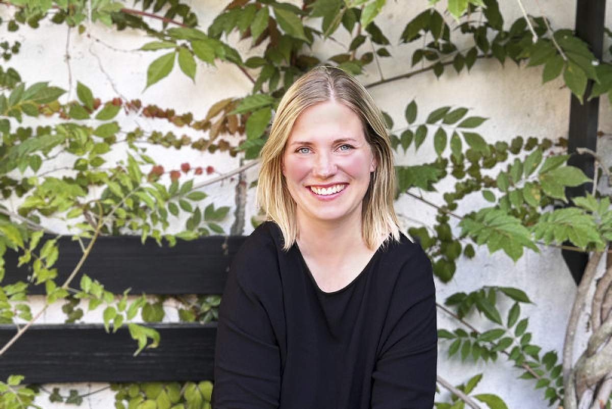 En kvinna med blont hår och svart tröja sitter framför en vit vägg med gröna rankor och ser in i kameran.