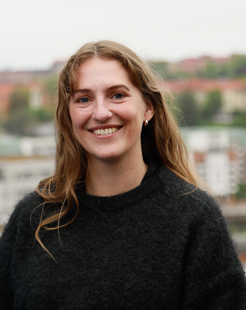 En kvinna med långt brunt hår och svart tröja ler mot kameran utomhus, med en suddig stadsmiljö i bakgrunden.