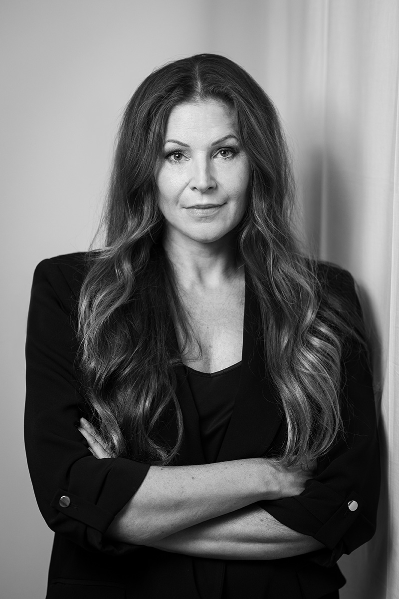 En kvinna med långt, vågigt hår och kavaj står med armarna i kors och tittar in i kameran på ett svartvitt foto.