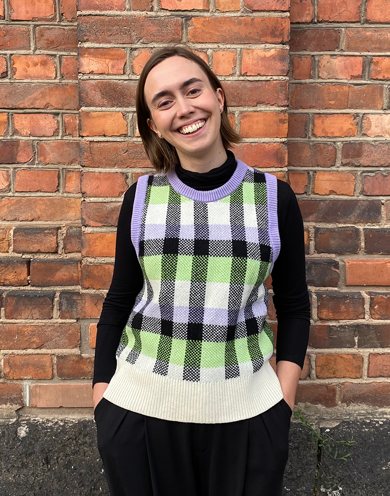 En person står framför en tegelvägg, klädd i en svart långärmad tröja och en grön, svart och vit rutig väst med lila kant.