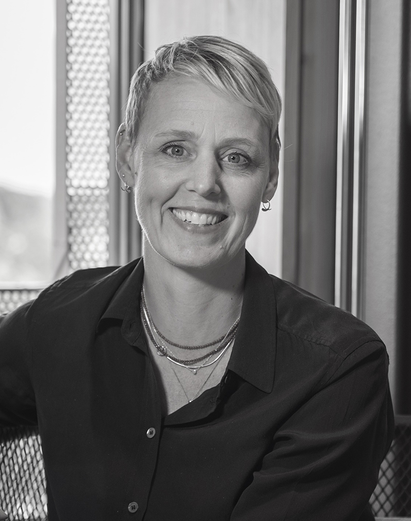 Svartvit porträttbild av en person med kort ljust hår, mörk skjorta och halsband, sittande inomhus vid ett fönster och ler.