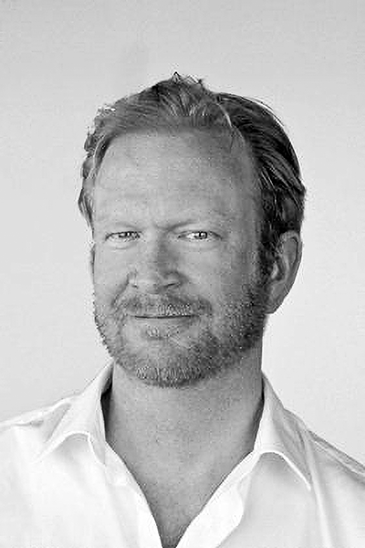 Porträtt i svartvitt av en medelålders man med kort hår och skägg, lätt leende, vit skjorta, vänd mot kameran.