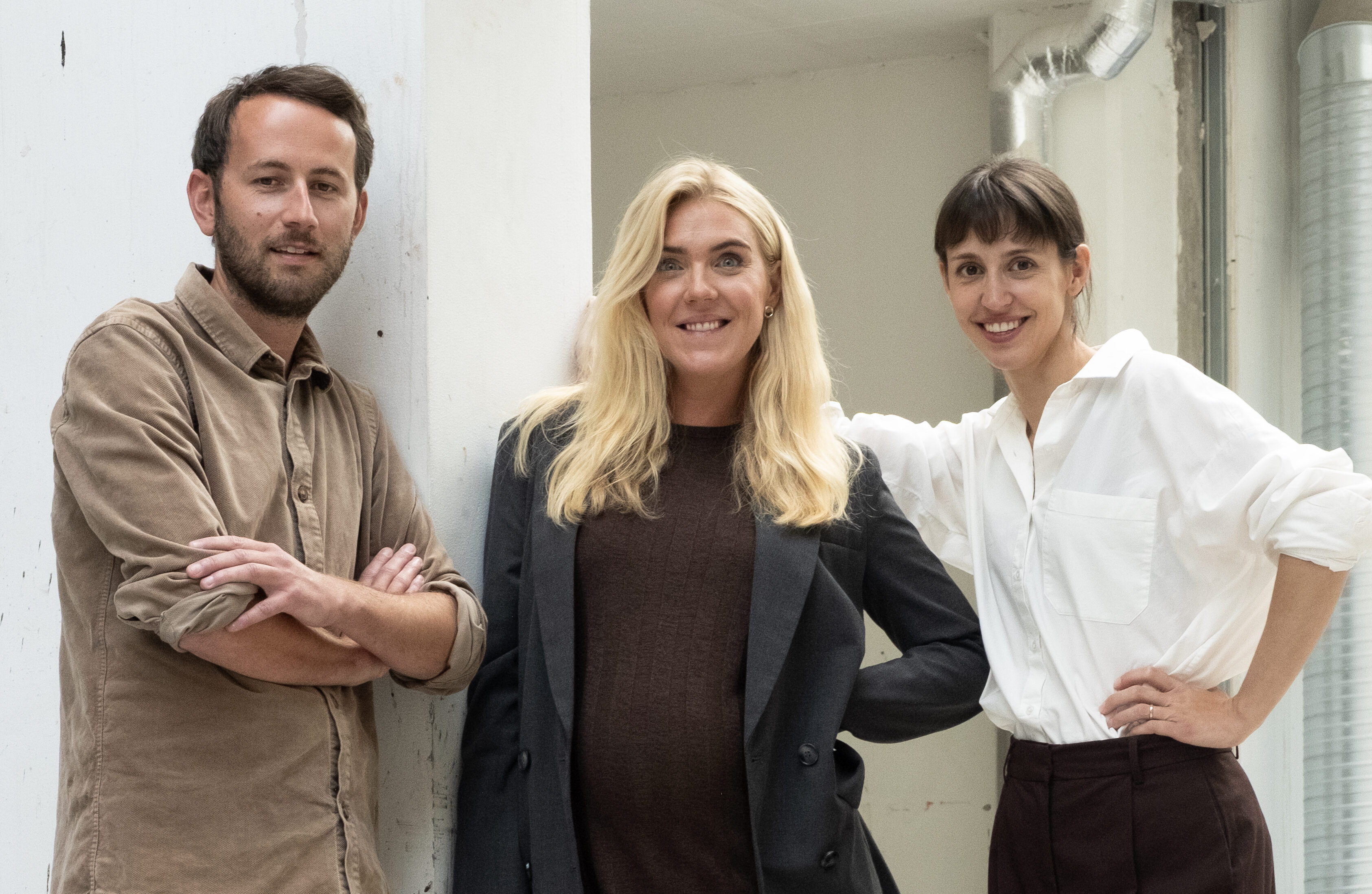 Tre personer står inomhus för ett foto. En lutar sig mot en vägg, två står intill varandra, alla ser mot kameran och ler.