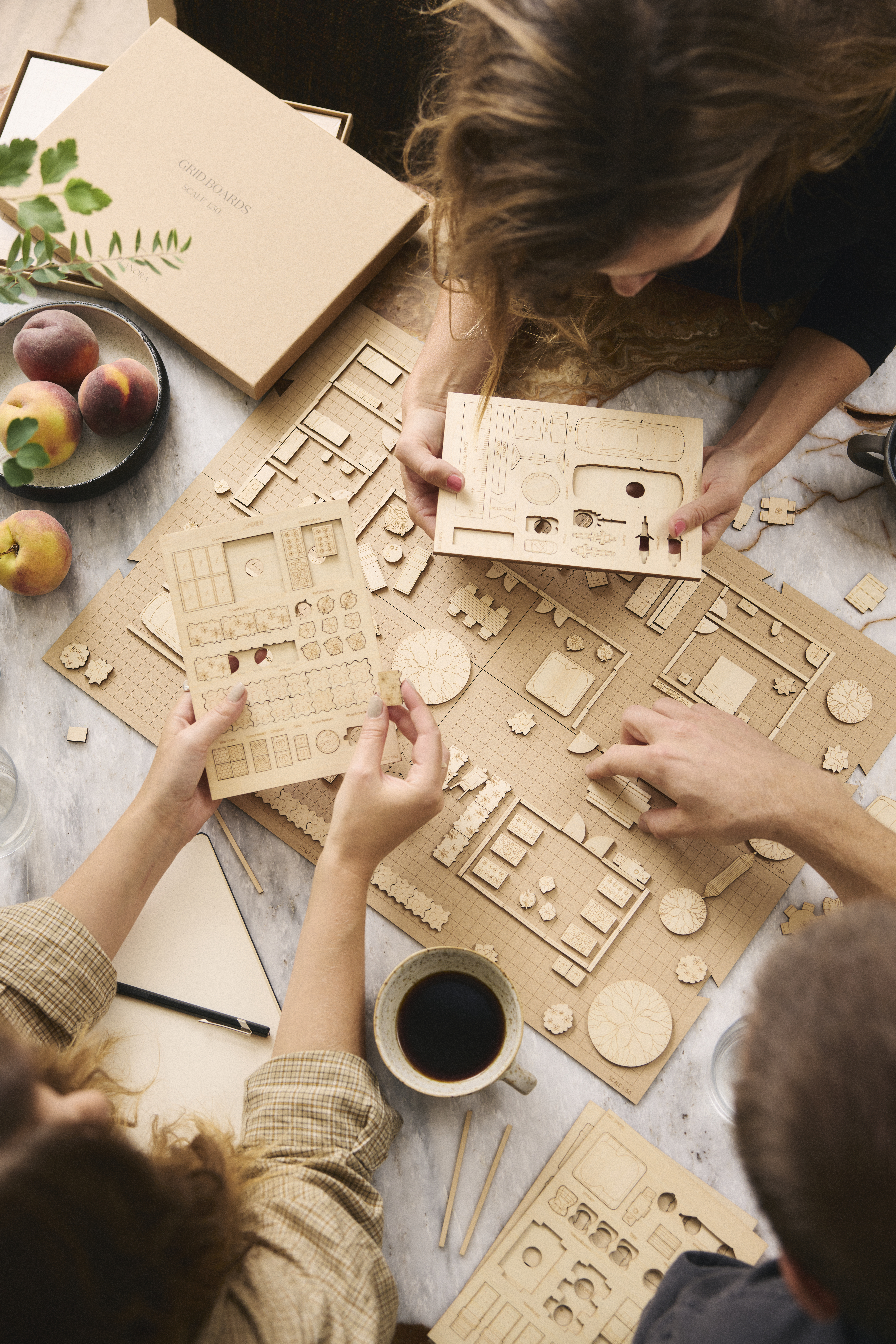 Tre personer sätter ihop en modell i trä vid ett bord med kaffe, persikor och delar utspridda, sedda uppifrån.
