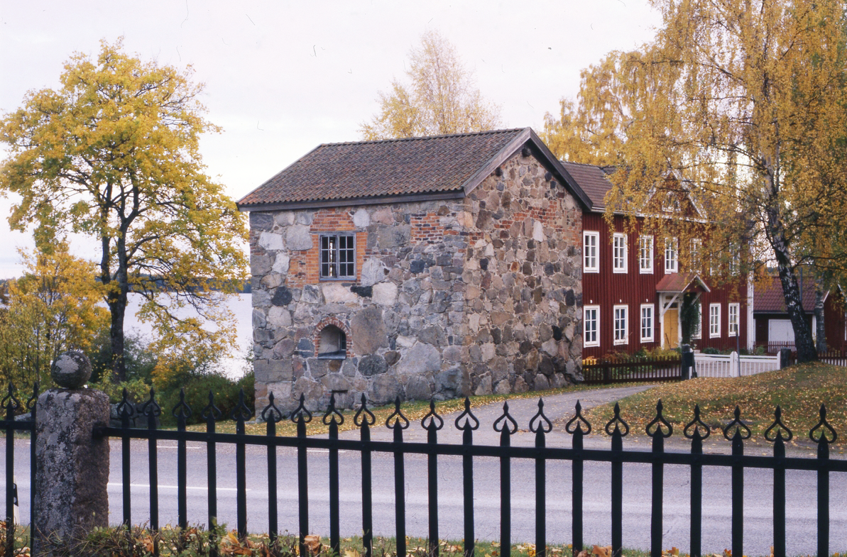 En sten- och tegelbyggnad står bredvid ett rött trähus, omgiven av träd och ett svart järnstaket vid en lugn väg.