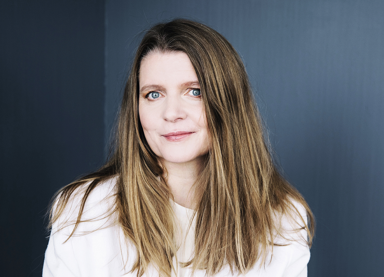 En kvinna med långt, rakt ljusbrunt hår och blå ögon bär en vit topp och står framför en mörkblå bakgrund.