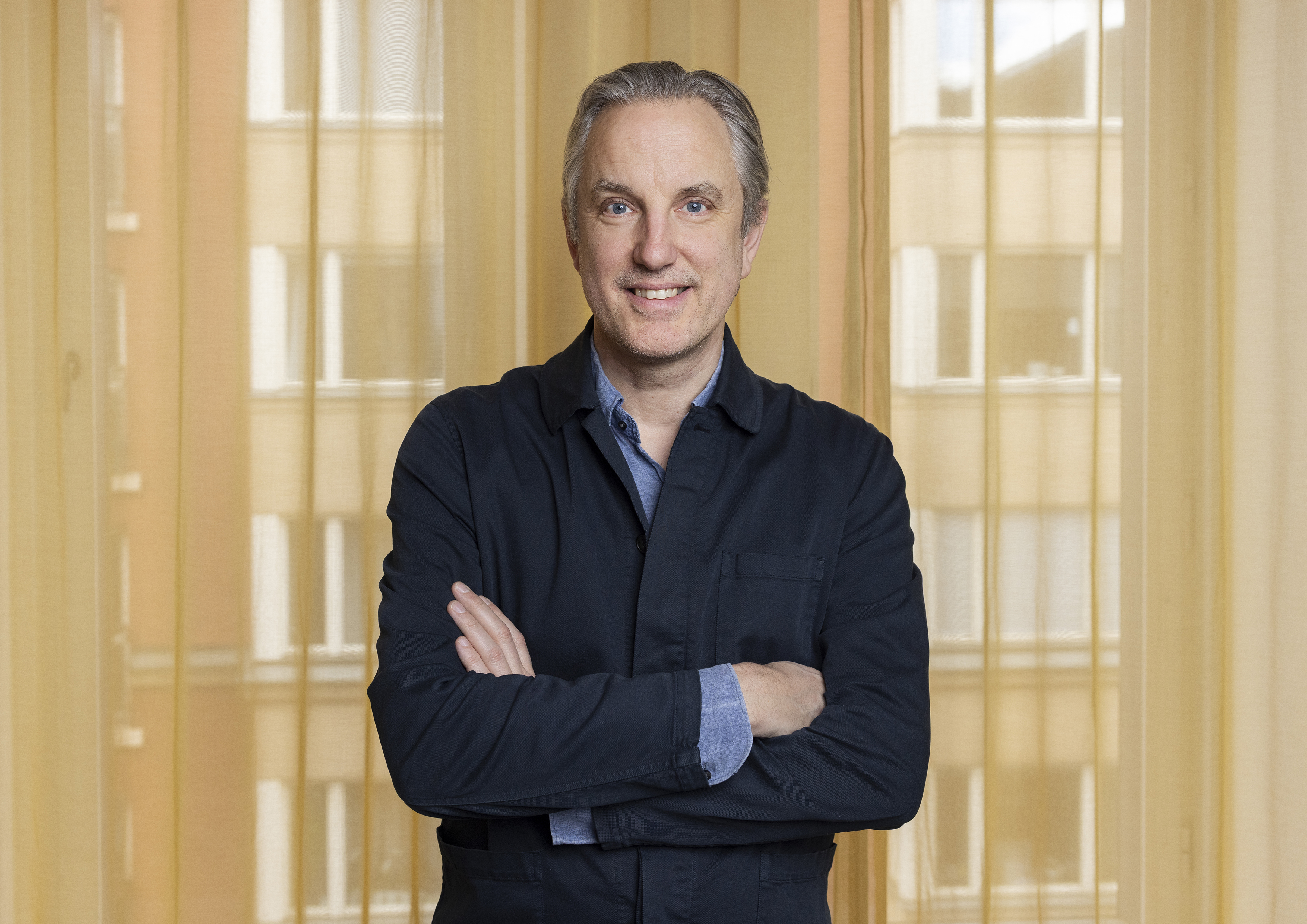En man med grått hår, i mörk jacka och blå skjorta, står med armarna i kors och ler framför gula gardiner och höga fönster.