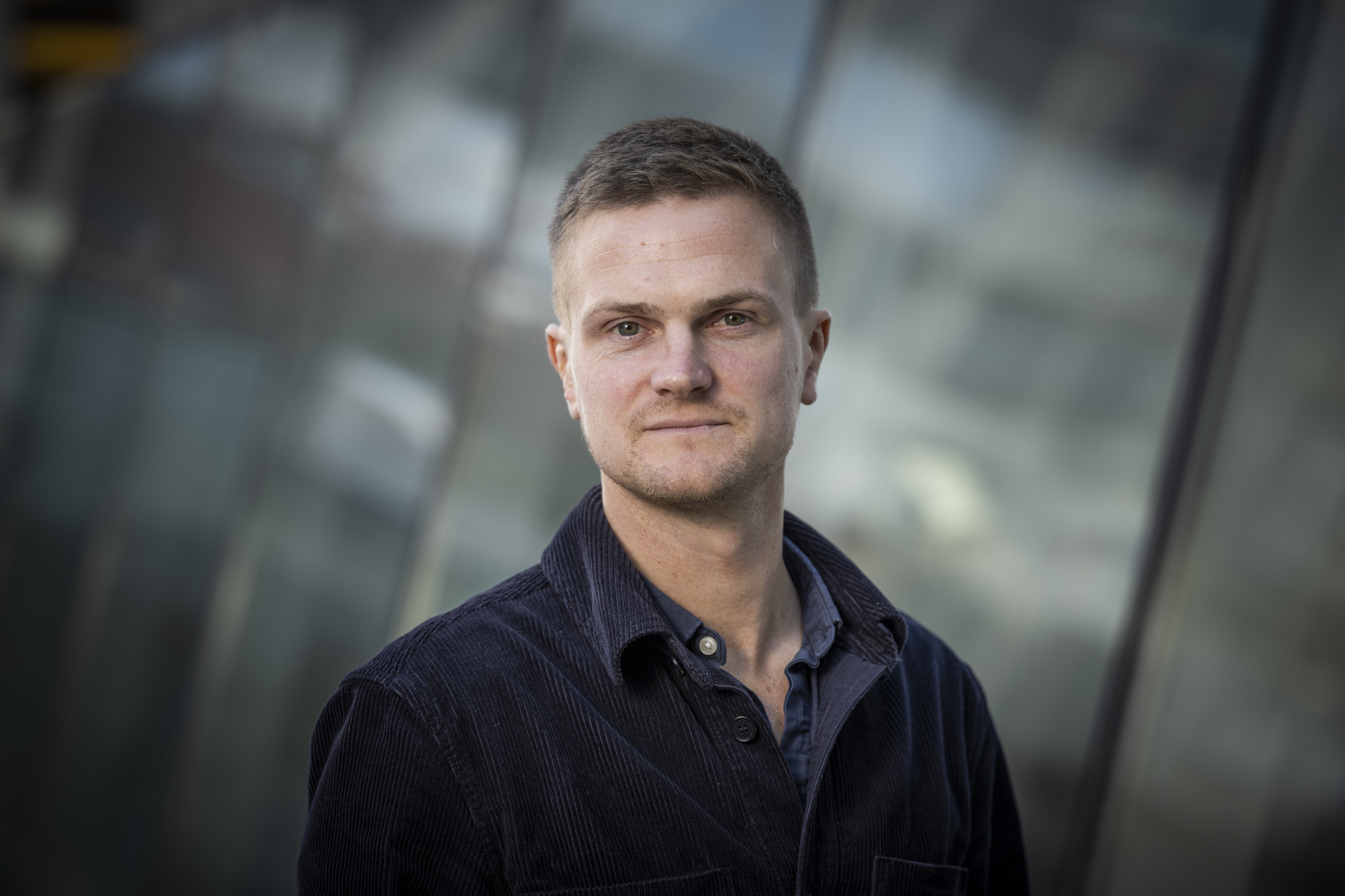 En man med kort hår och neutralt ansiktsuttryck står utomhus framför en glasbyggnads fasad.