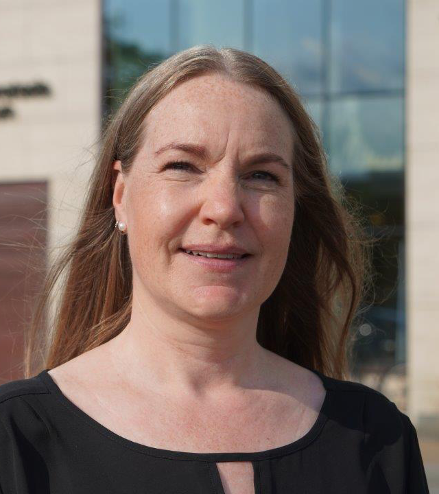 En kvinna med långt ljusbrunt hår och svart topp står utomhus framför en byggnad med stora glasfönster.