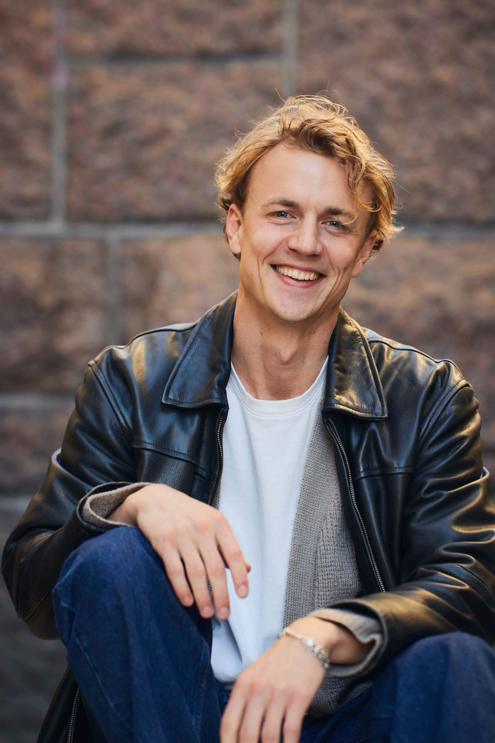 En ung man med blont hår, klädd i svart skinnjacka och vit skjorta, sitter och ler framför en stenmur.