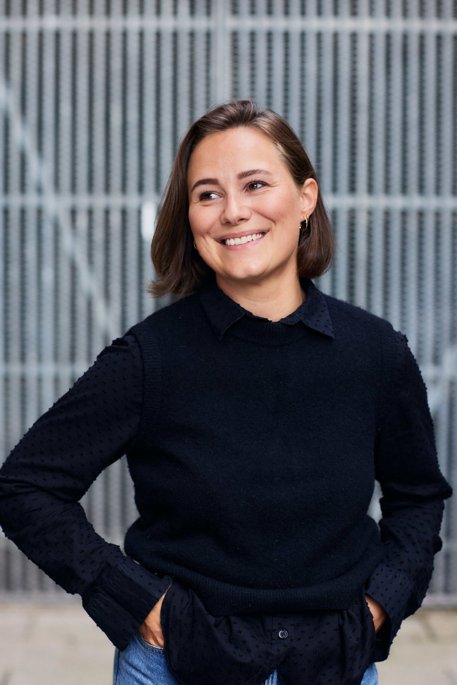 En kvinna med kort brunt hår, svart tröja och blå jeans står framför ett staket med händerna i fickorna och ler.