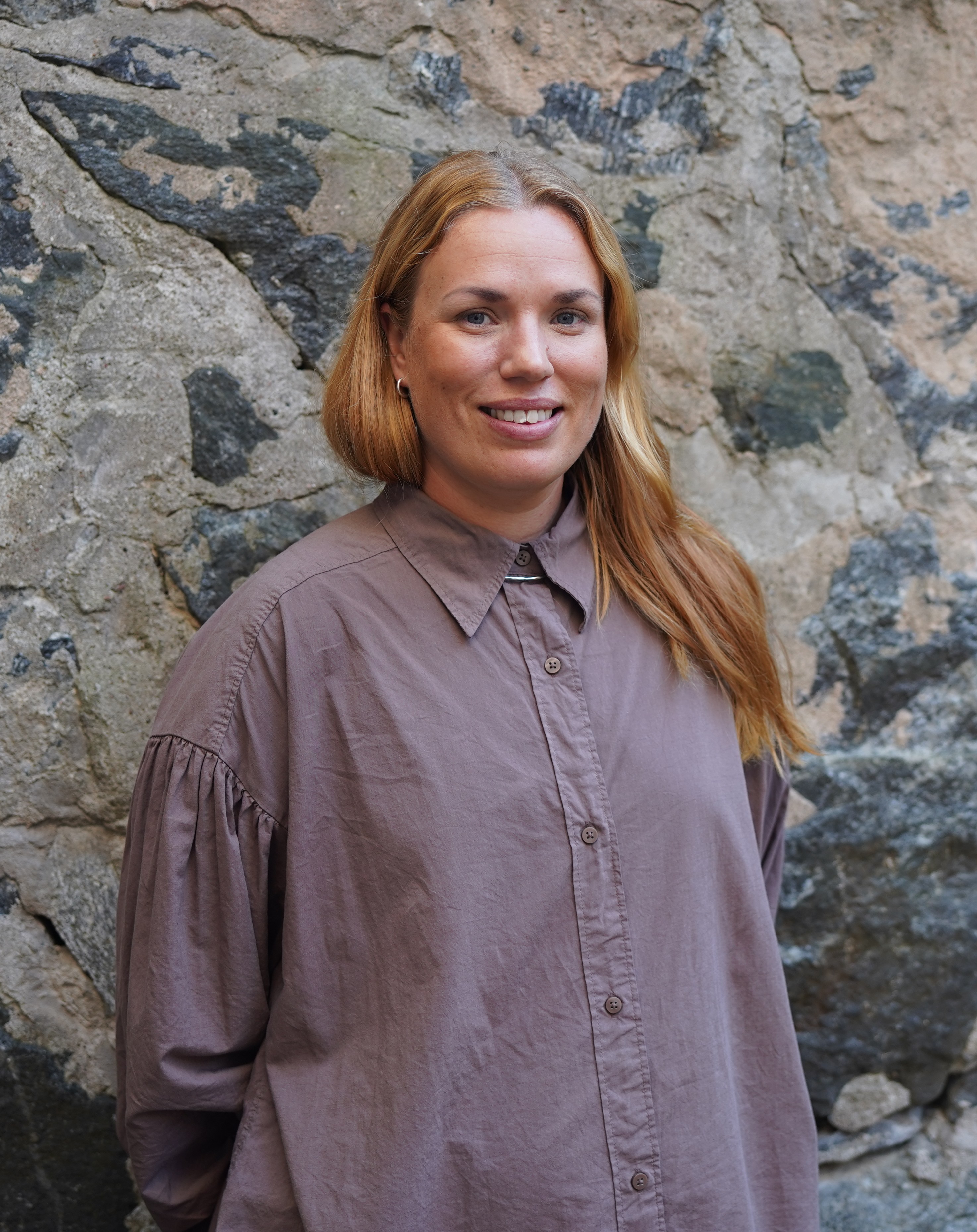En kvinna med långt ljusbrunt hår och brun skjorta står framför en stenmur och ler mot kameran.