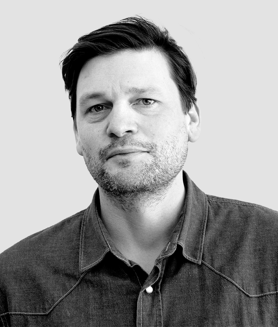 En man med kort mörkt hår och lätt skägg bär en jeansskjorta och tittar mot kameran framför en ljus bakgrund.