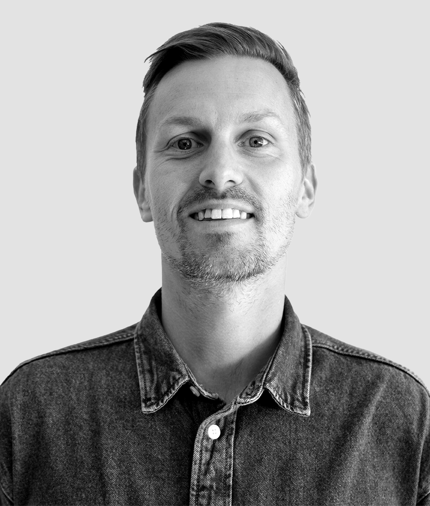 En man med kort hår och lätt skäggväxt, iklädd en knäppt jeansskjorta, vänd mot kameran mot en ljus bakgrund.