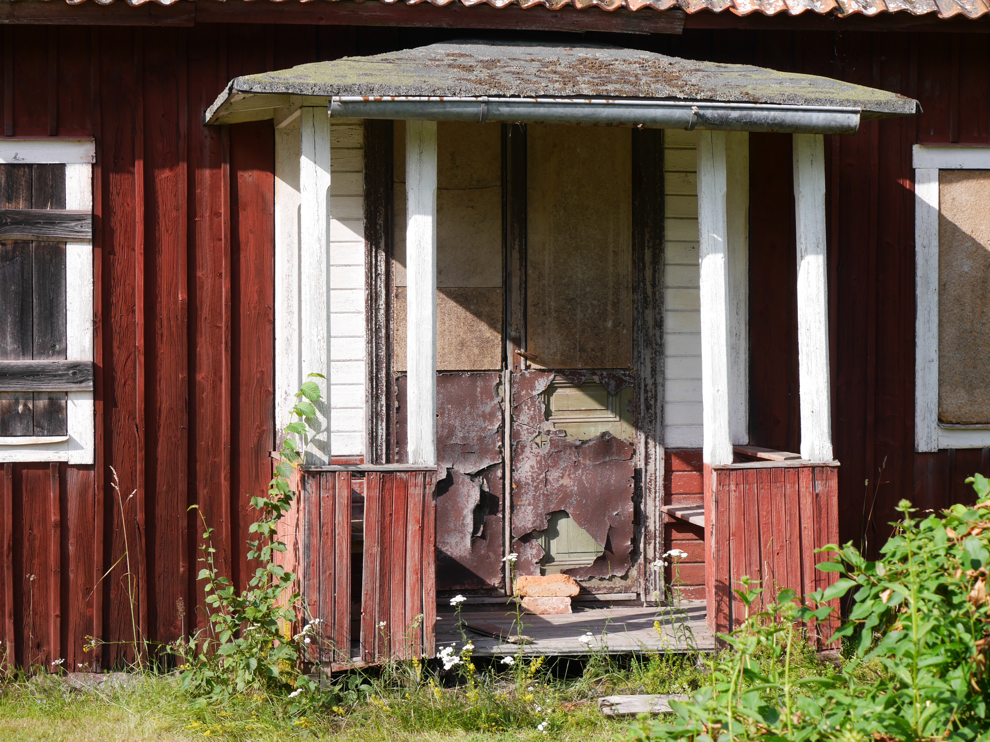 En närbild på en övergiven veranda med röd färg som flagnar, trasigt trä och växter framför en byggnad.