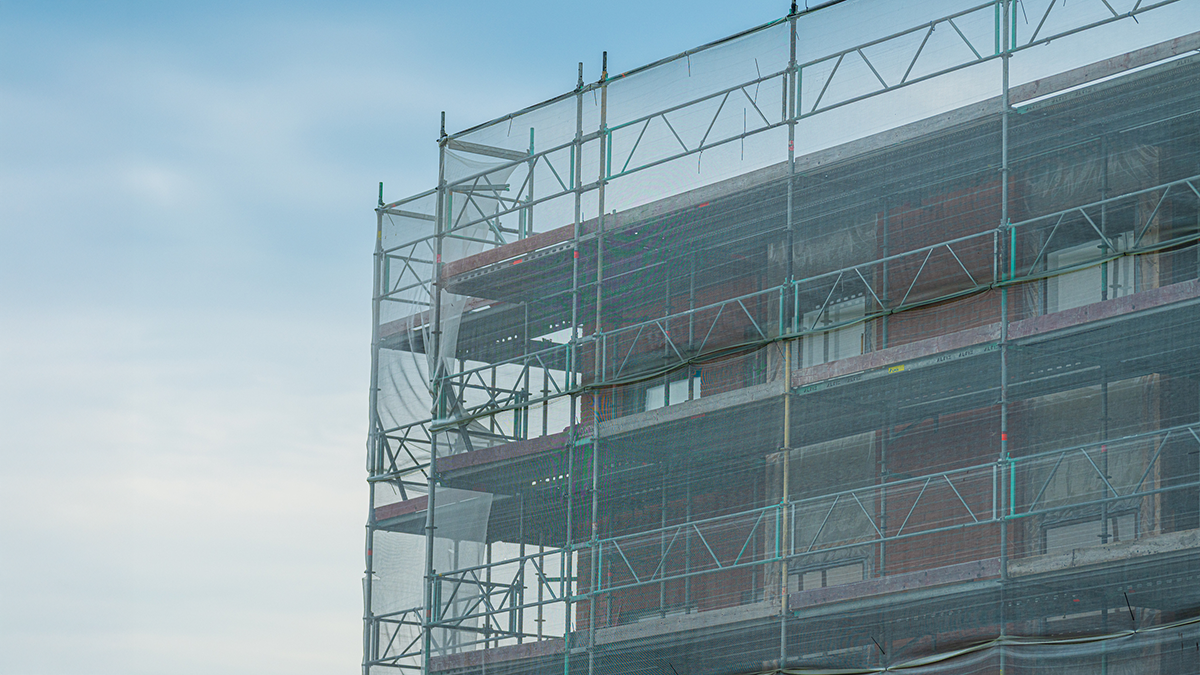 En byggnad under uppförande är täckt av ställningar och grönt nätväv mot en blå himmel.