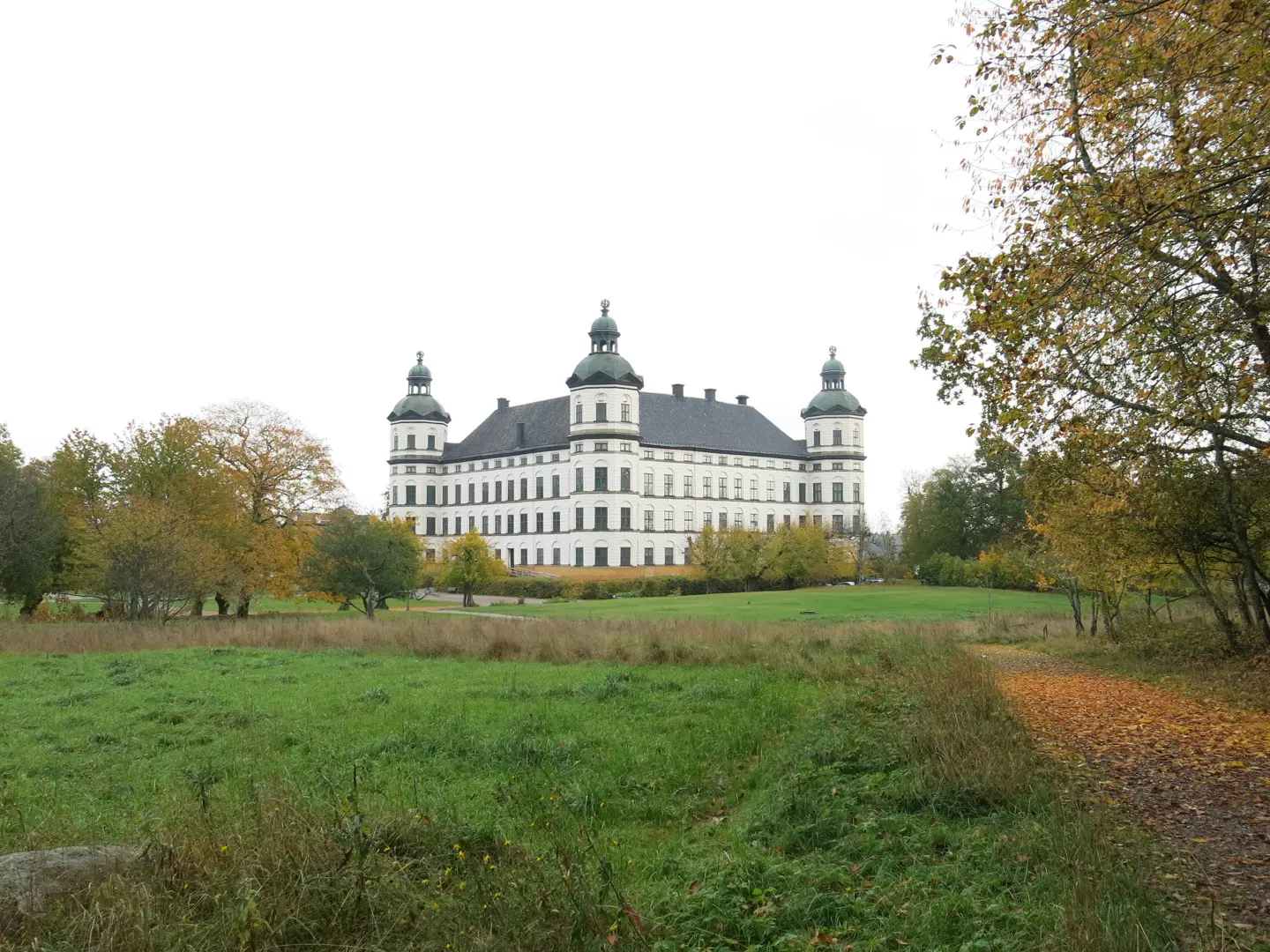 Ett vitt historiskt byggnad med fyra kupoltorn står bakom en gräsplan och är omgiven av träd i höst.
