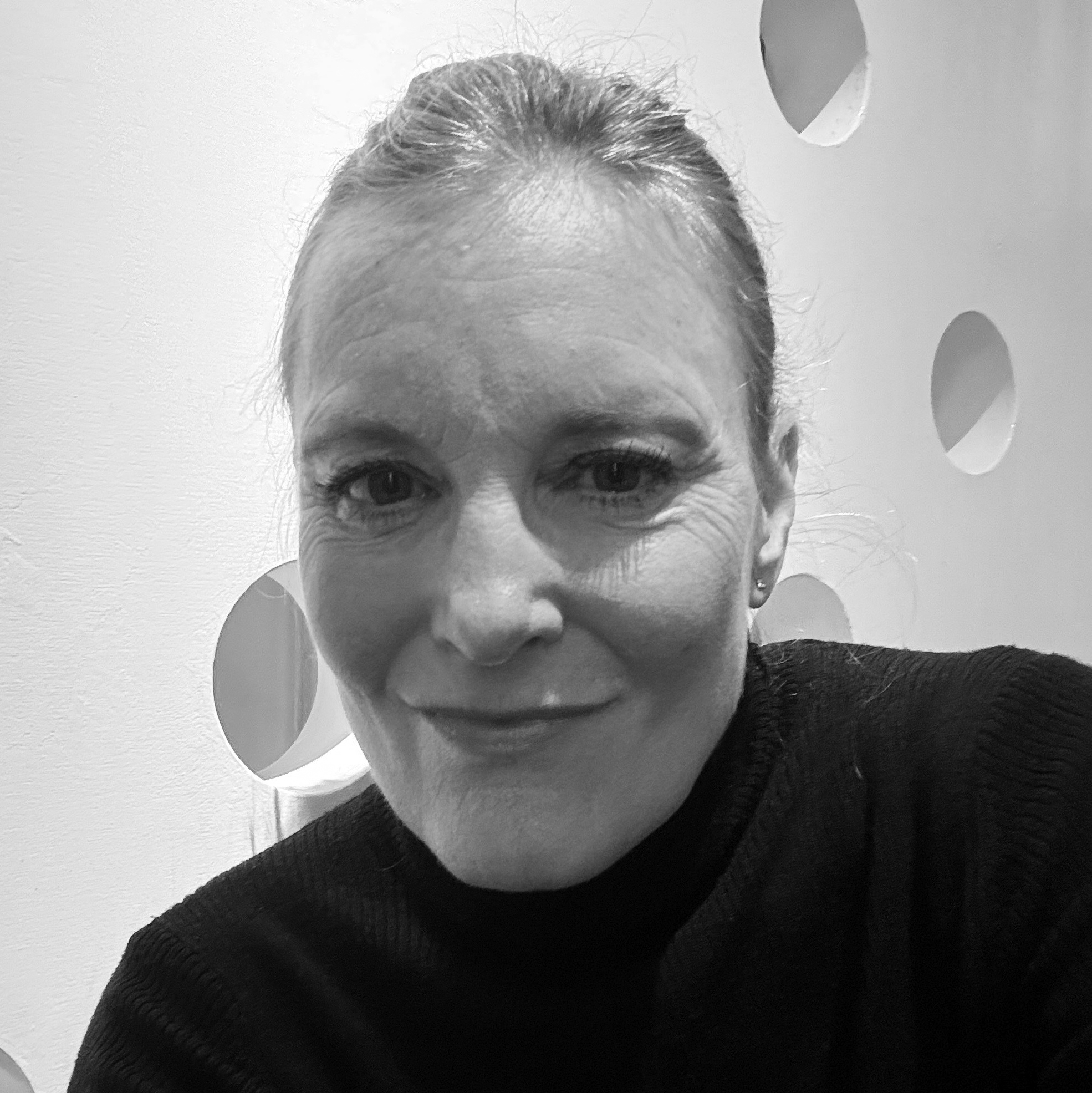 En kvinna med ljust hår uppsatt och mörk topp ler mot kameran framför en vit vägg med runda utskärningar. Svartvit foto.