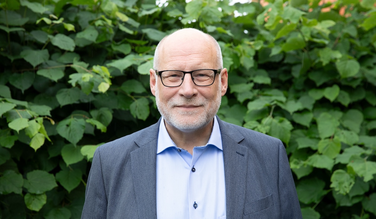 En man med glasögon och skägg, klädd i blå skjorta och grå kavaj, står framför gröna buskar.