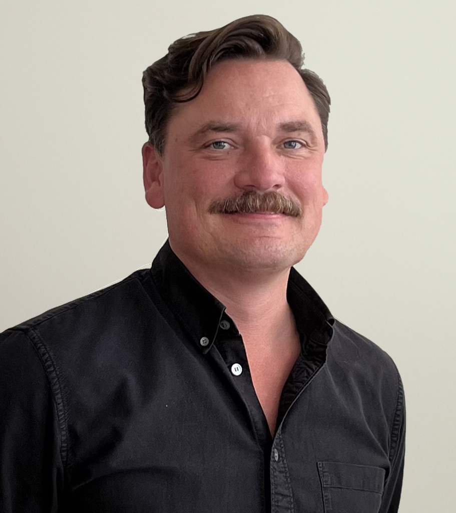 En man med brunt hår och mustasch har på sig en svart skjorta och ler mot kameran framför en ljus bakgrund.