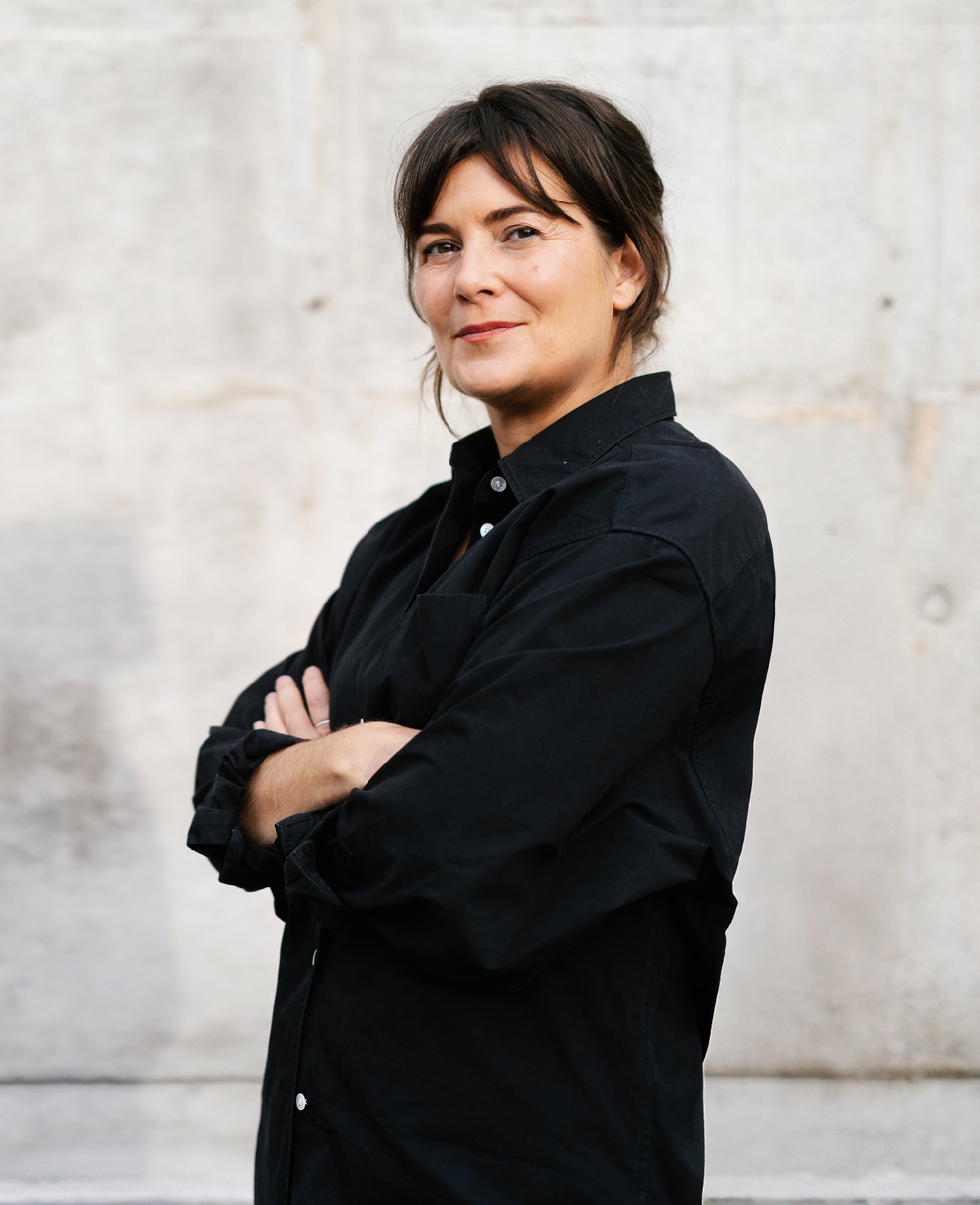 En kvinna med mörkt hår samlat i en låg knut står framför en ljus betongvägg, iförd svart skjorta med armarna i kors.