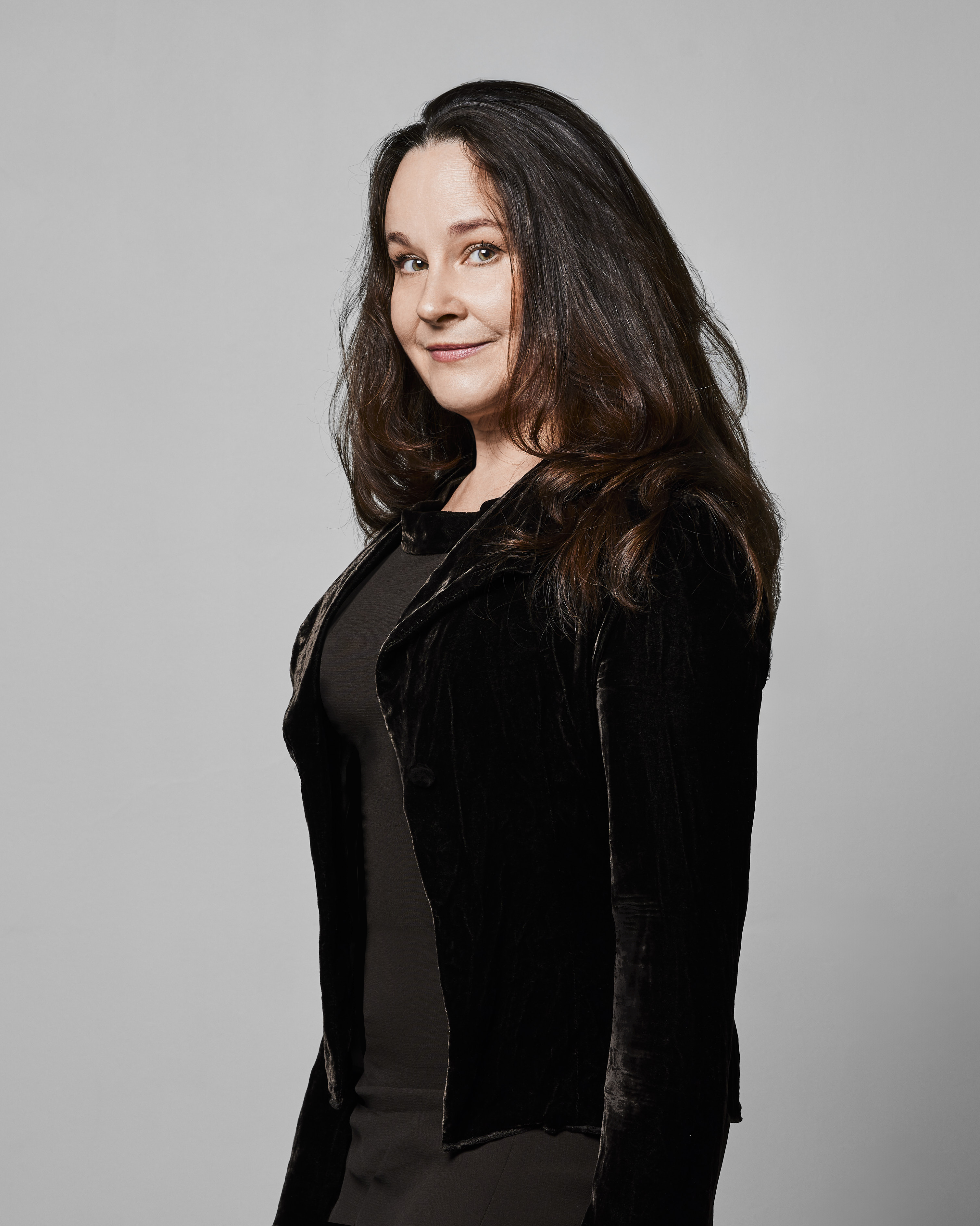 Kvinna med långt mörkt hår i svart jacka och klänning står och tittar mot kameran mot en ljusgrå bakgrund.