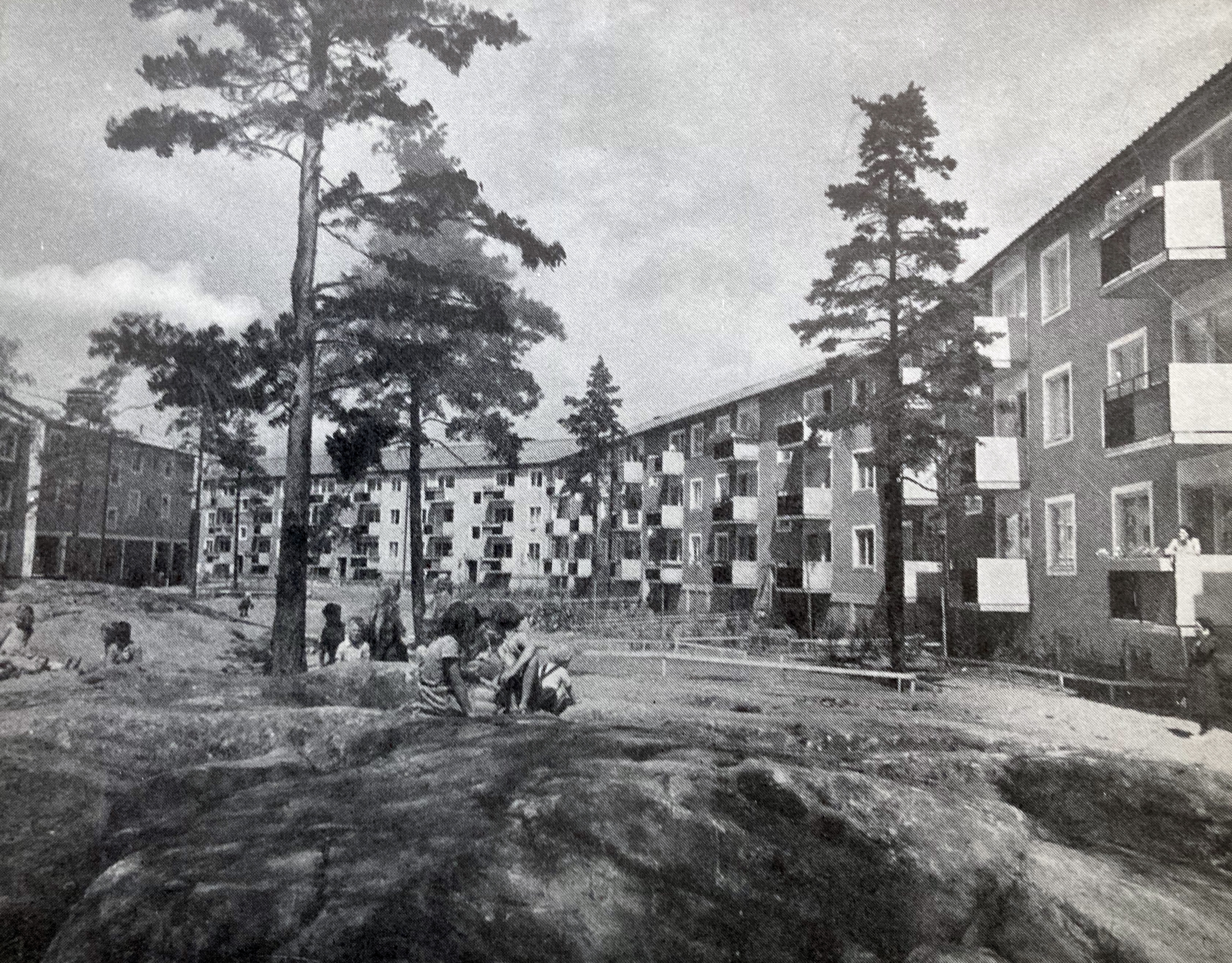Svartvit bild av barn som leker på stenig mark framför flerbostadshus från mitten av 1900-talet med tallar runtom.