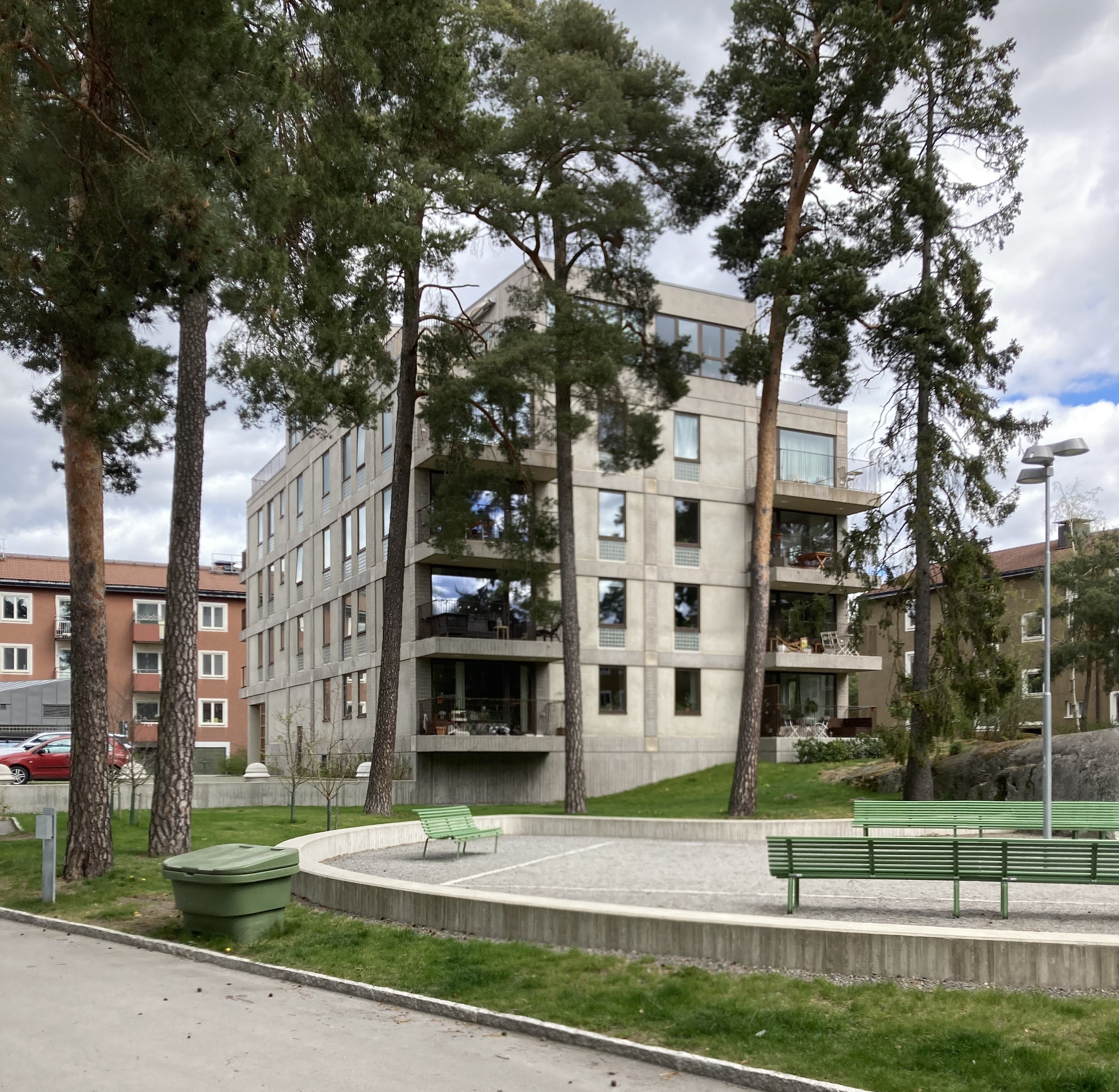 Ett fyravåningshus i betong står bakom tallar, med gröna bänkar och en cirkelformad stenlagd yta framför.