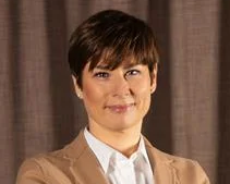 En person med kort brunt hår och en beige kavaj samt vit skjorta står framför ett brunt draperi.