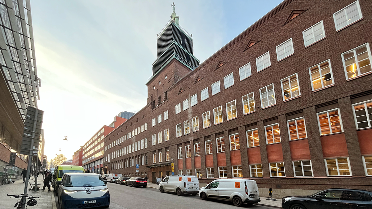 En gata med en tegelbyggnad med många fönster och ett torn, bilar och skåpbilar parkerade på båda sidor.