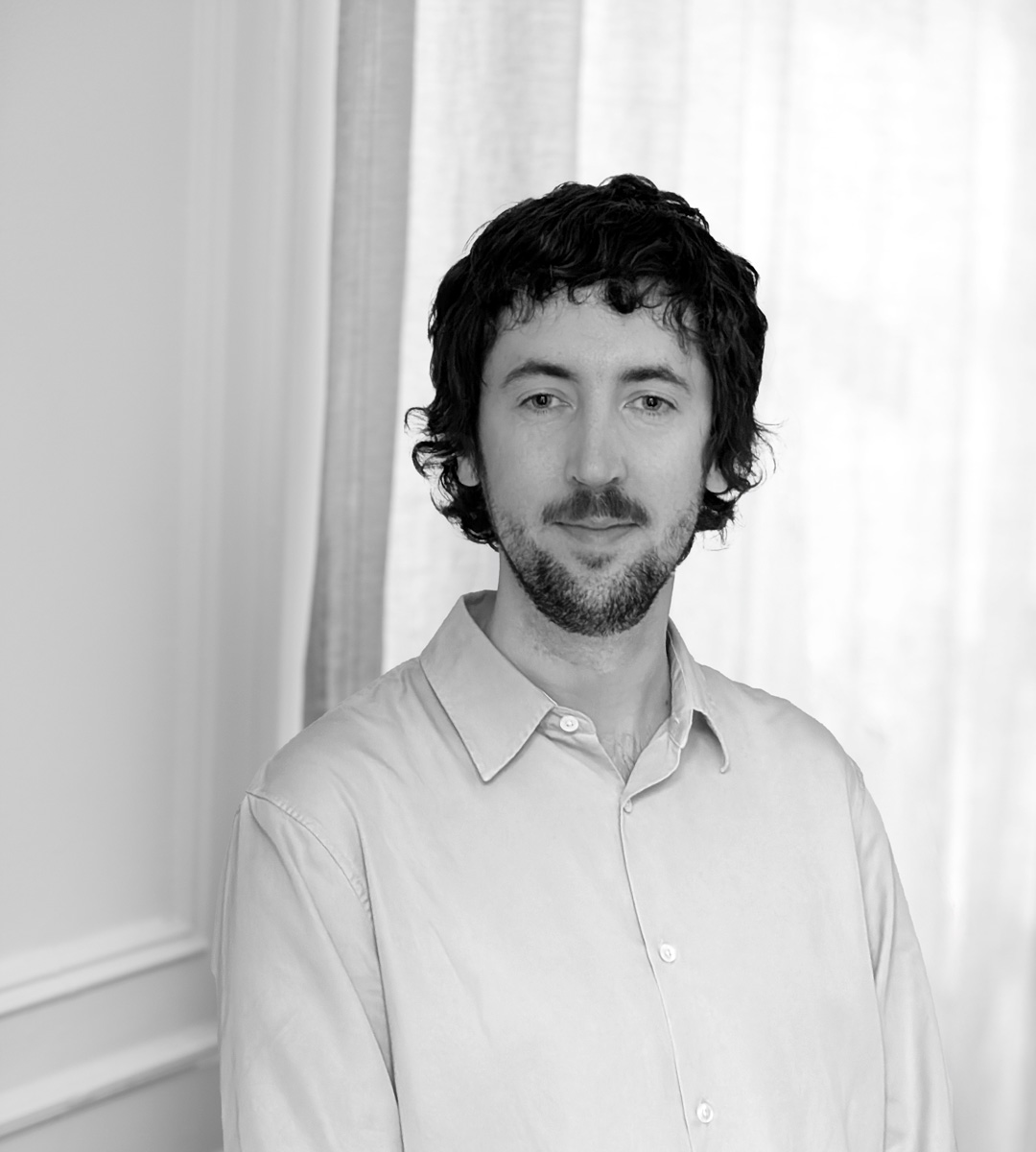 En man med mörkt lockigt hår och skägg har på sig en ljus skjorta och står framför ljusa gardiner, tittar mot kameran.