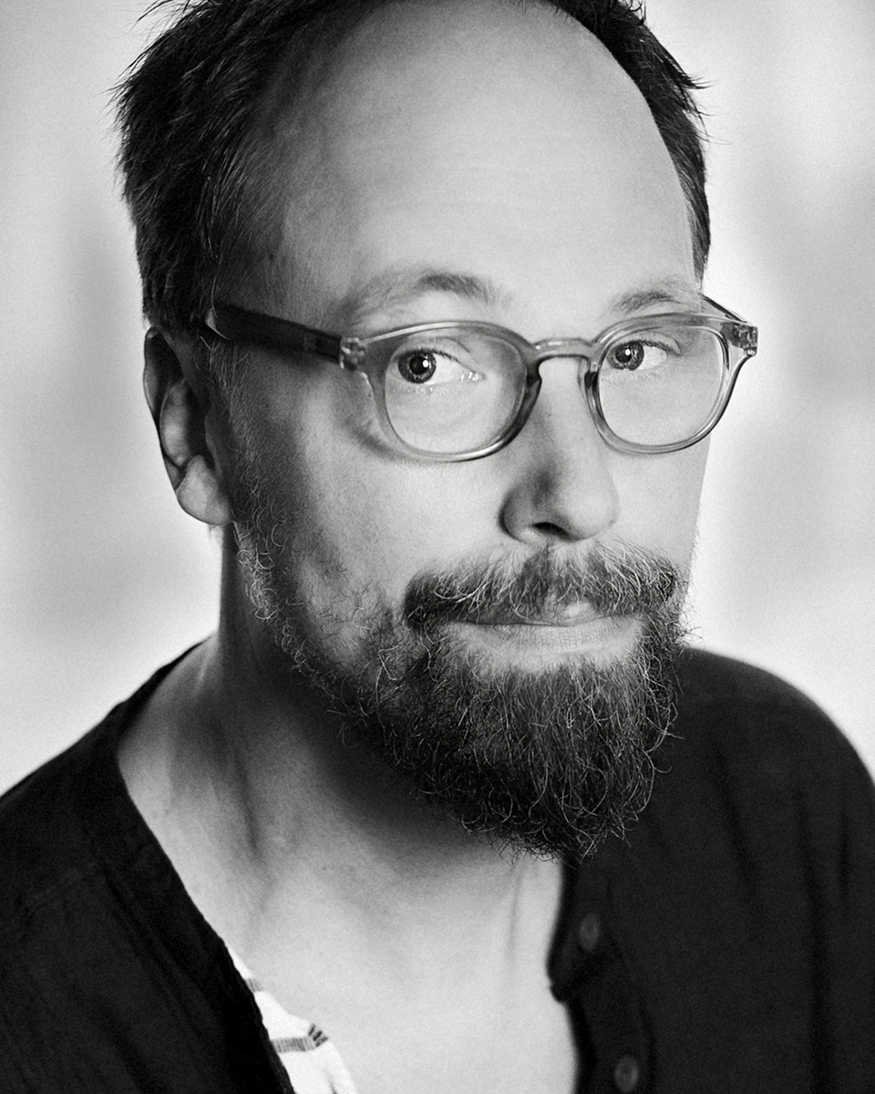 En man med glasögon och skägg ser mot kameran, har en mörk skjorta i ett svartvitt porträtt med en ljus bakgrund.