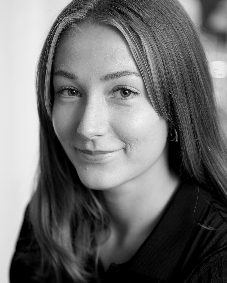 Porträtt i svartvitt av en ung kvinna med långt hår och mörk tröja som tittar mot kameran och ler lite.