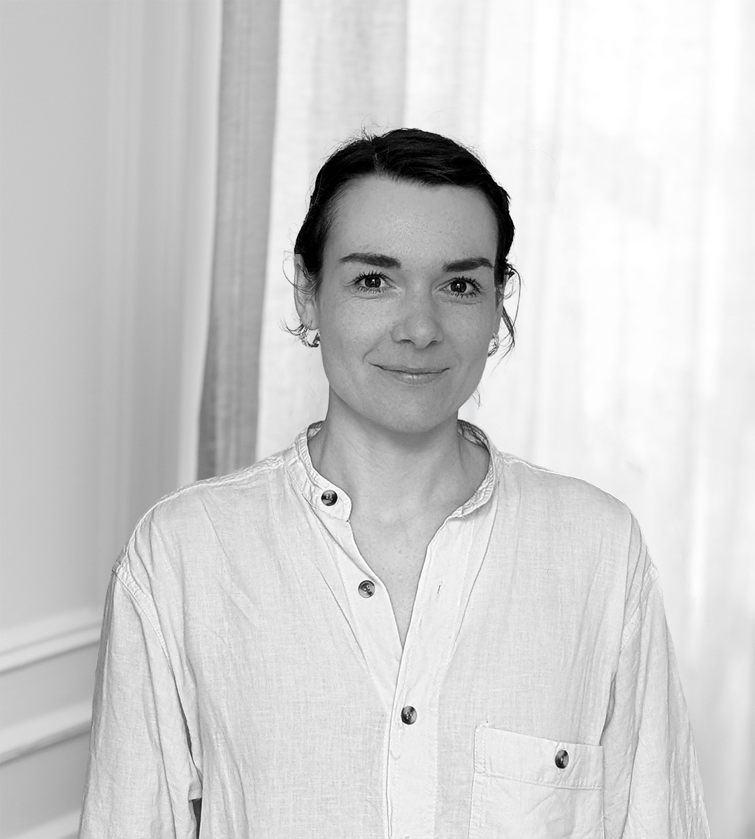 En kvinna med mörkt hår i en lös knut, iklädd en ljus skjorta med knappar, står inomhus framför ett draperi.