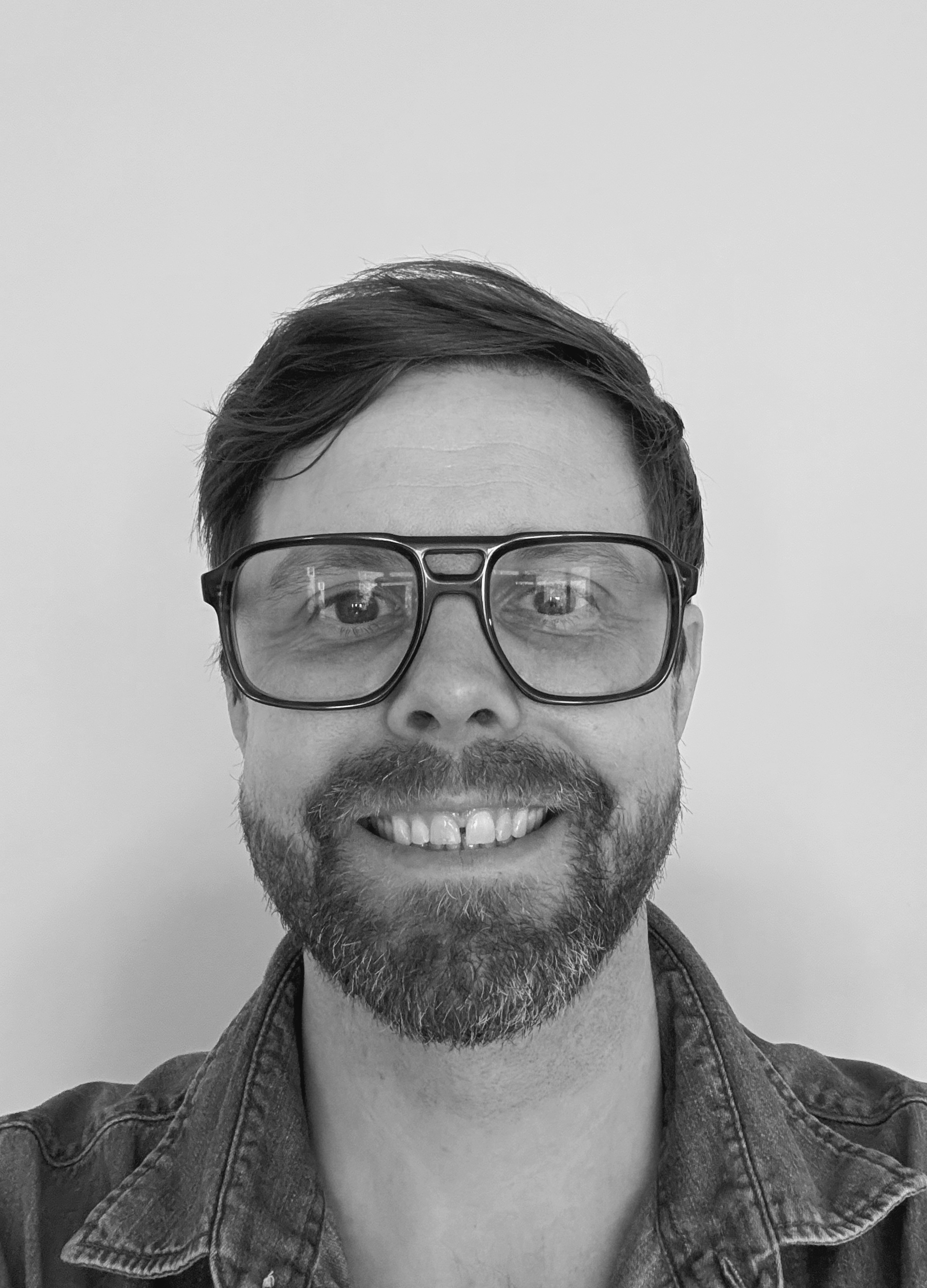En man med kort hår, skägg och stora glasögon tittar mot kameran. Han har på sig en jeansskjorta och står framför en enkel bakgrund.