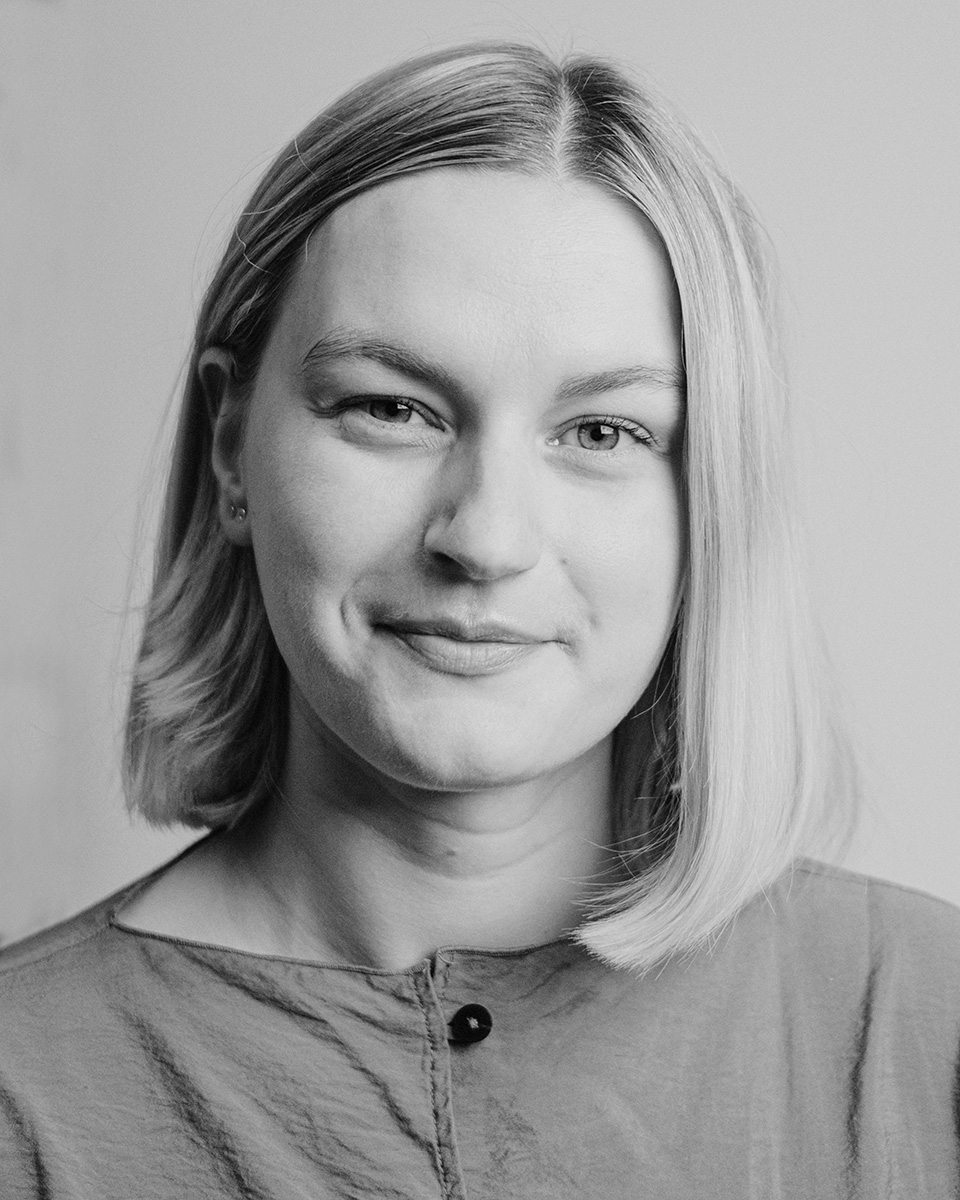 En kvinna med rakt, axellångt blont hår och neutralt ansiktsuttryck bär en knäppt skjorta framför en enkel bakgrund. Svartvit bild.