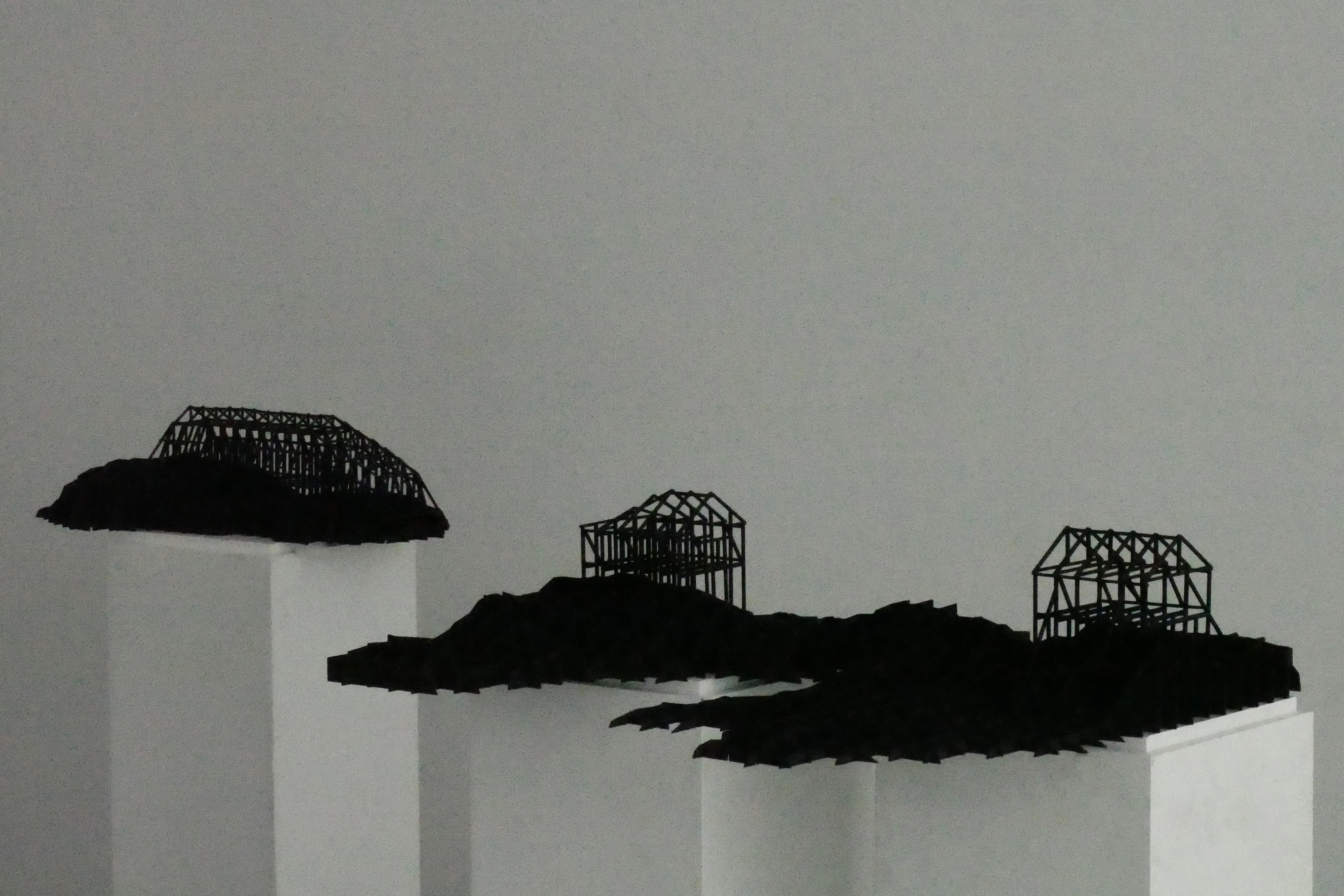 Tre svarta trådhusmodeller står på vita piedestaler, placerade på oregelbundna svarta ytor mot en grå bakgrund.