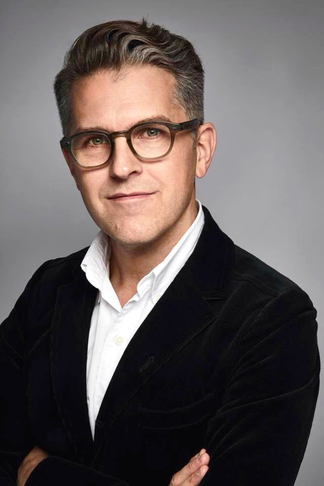 En man med kort hår med grå inslag och glasögon har vit skjorta och mörk kavaj, korsar armarna mot en grå bakgrund.