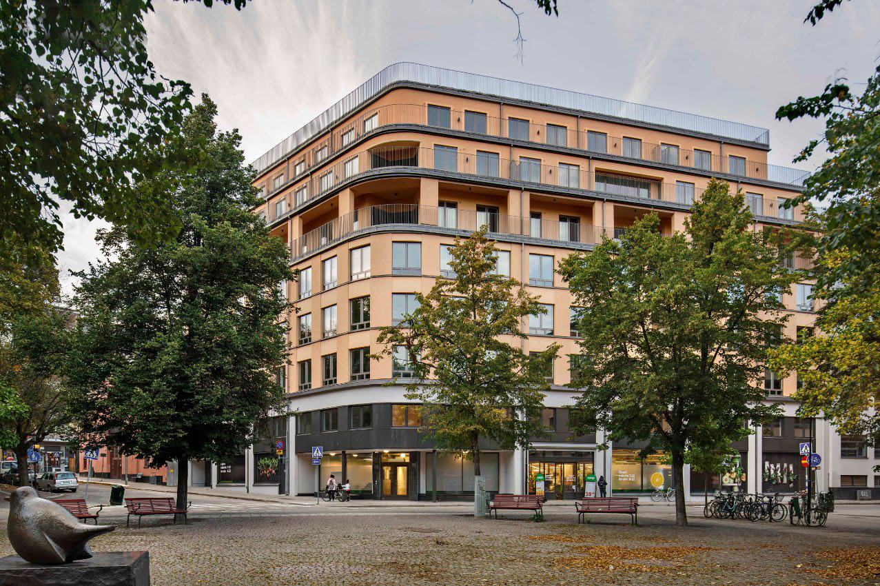 Ett flerbostadshus med balkonger står bakom träd och ett torg med bänkar och en skulptur i förgrunden.