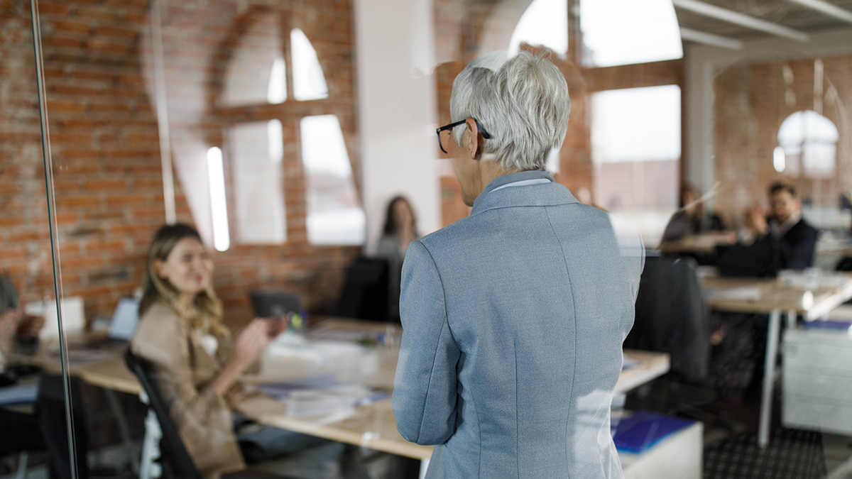 En person med kort grått hår står och pratar till en grupp människor som sitter vid skrivbord i ett kontor med tegelväggar och stora fönster.