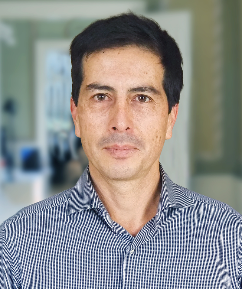 En man med kort mörkt hår och en rutig skjort står inomhus med en suddig bakgrund.